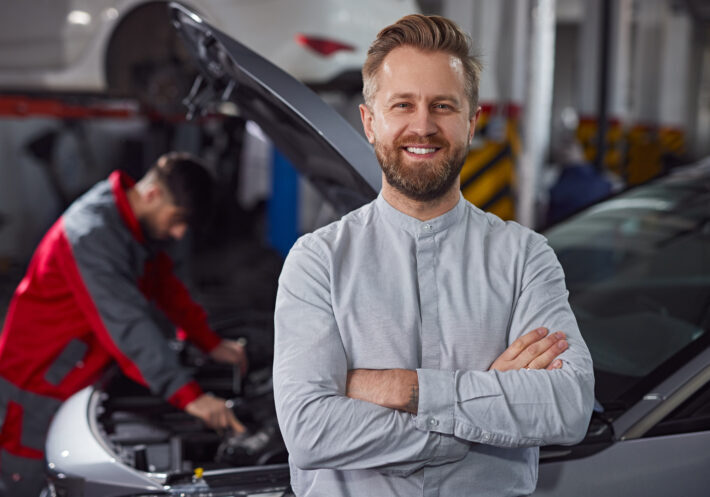 Confident customer near mechanic fixing car Positive bearded man with crossed arms smiling and looking at camera while standing near technician repairing vehicle in garage